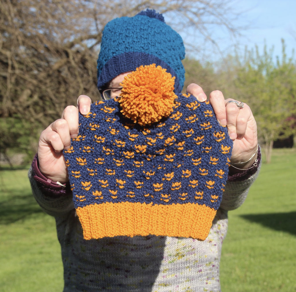 A woman wearing a blue hat stands in her garden, hold a small handknit hat in navy with bright yellow brim, accent stitches and a pompom.