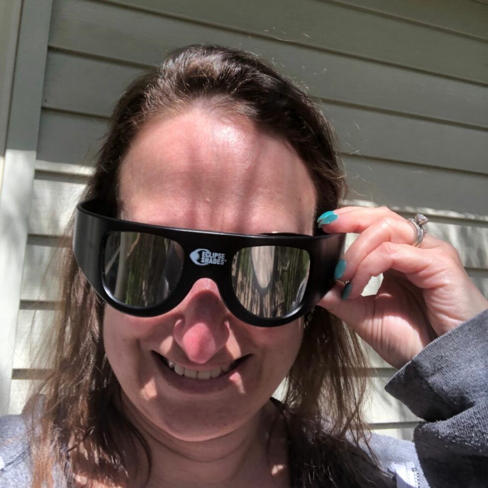 A woman wearing mirrored eclipse glasses smiles at the camera.