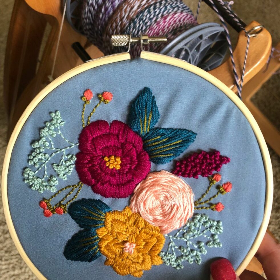 Embroidery flowers in red, peach and gold against a blue background in a hoop.