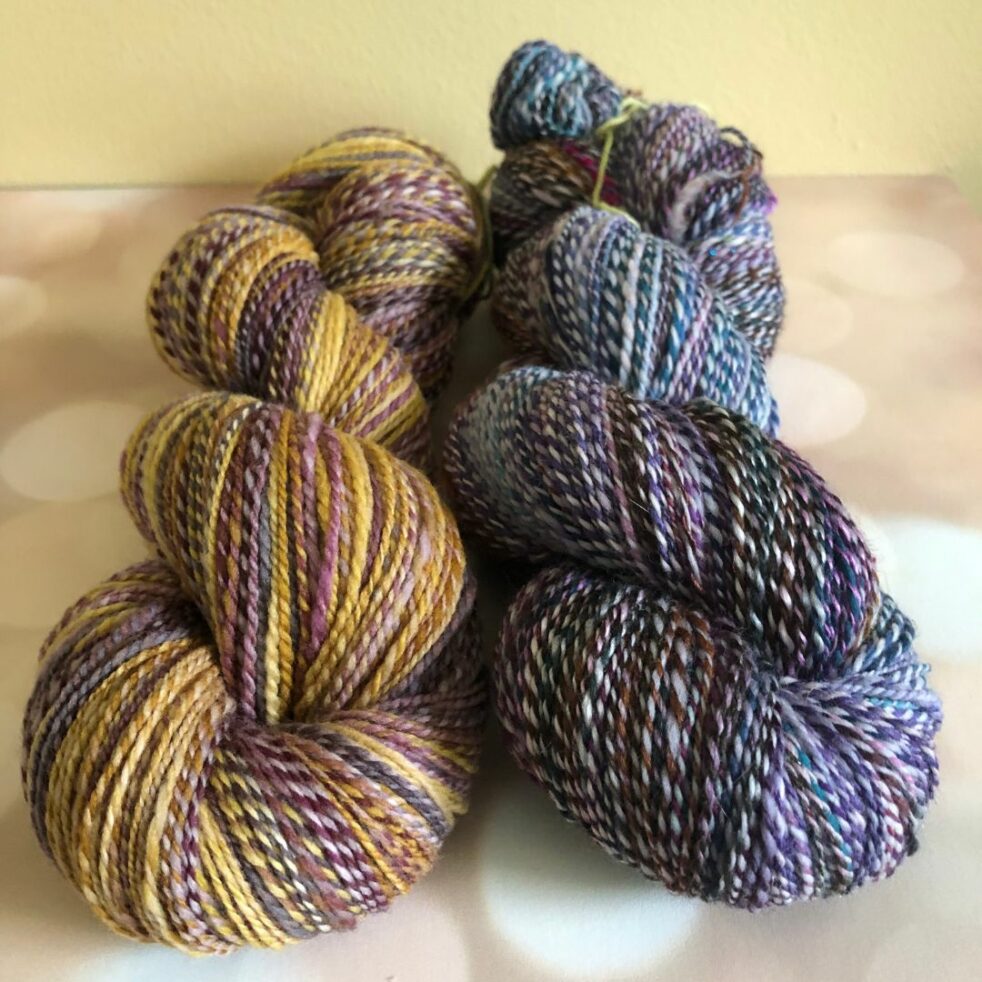 Two skeins of handspun yarn, with barberpole plies. On the left, the skein is gold, mauve and pink. On the right the skein is purple and lavender marled tones.